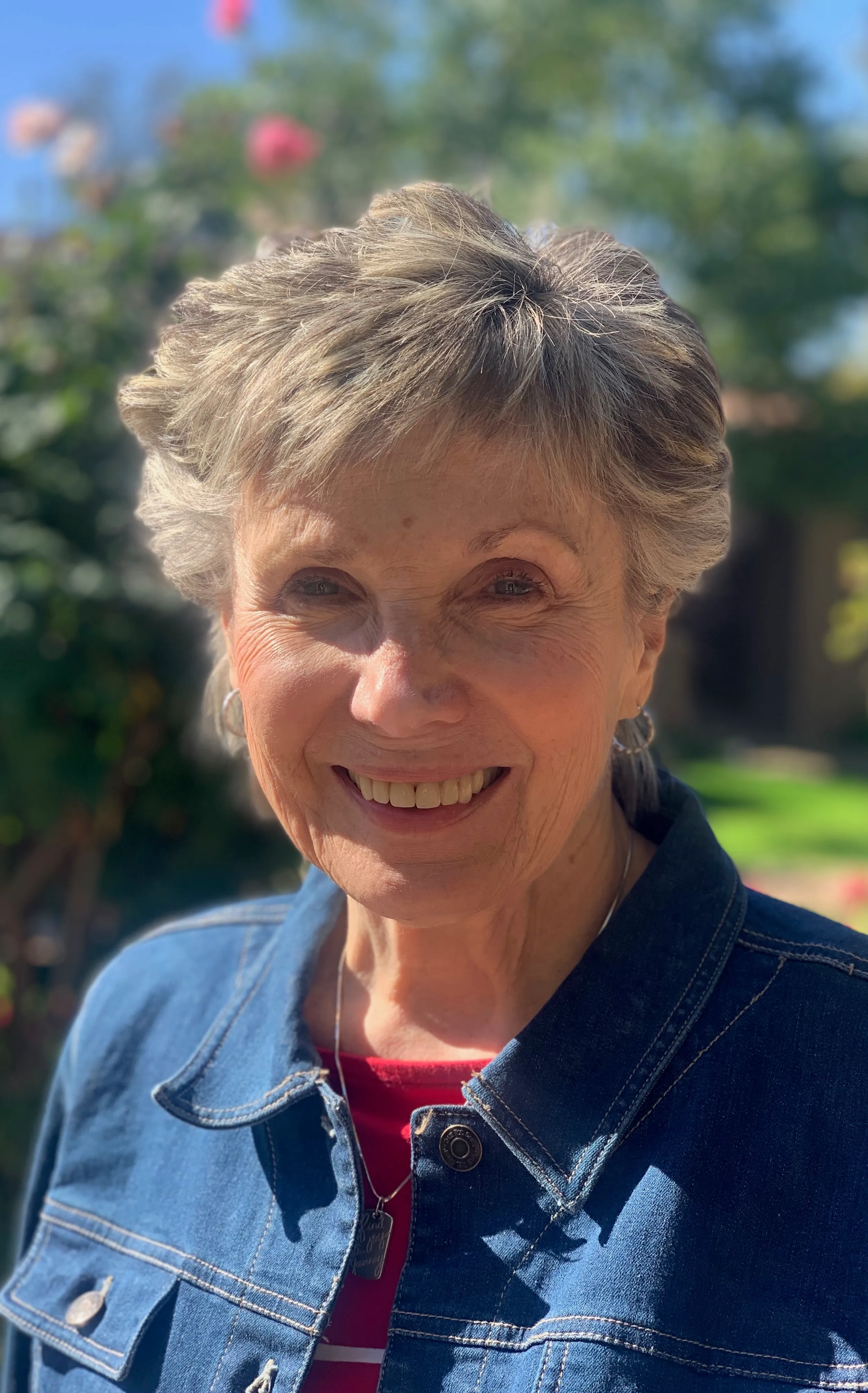 Mary Kay smiling outdoors in a denim jacket with a sunlit garden behind her